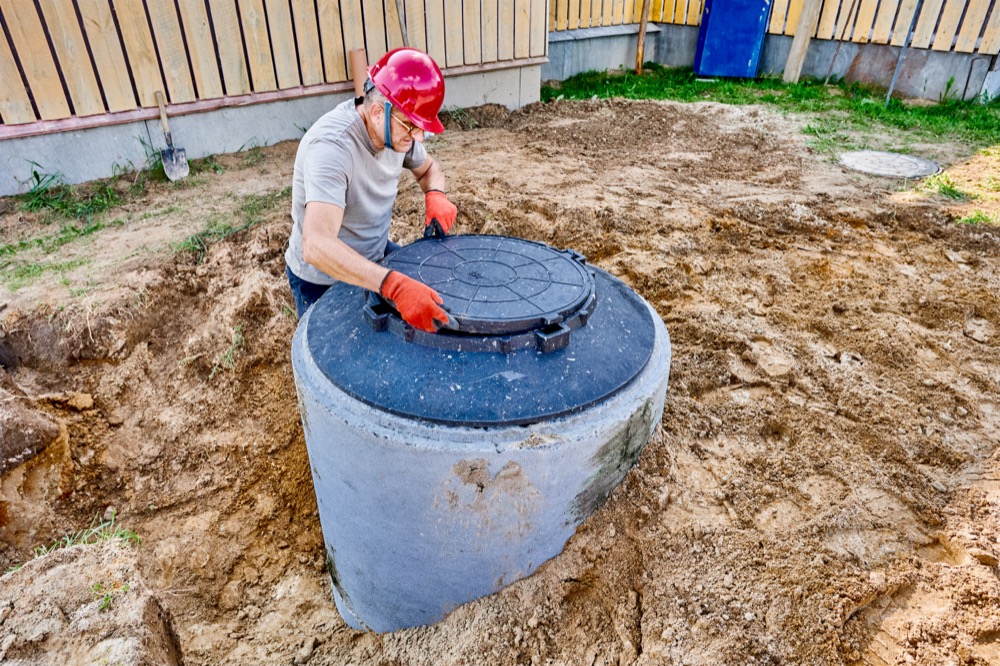 Construction worker in a hard hat lowering a circular concrete septic tank lid into place at a residential site.